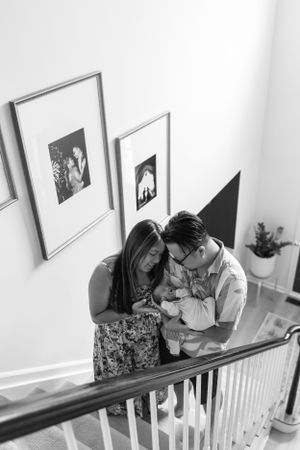 Family shares an intimate moment with newborn baby on stairs near framed photos.