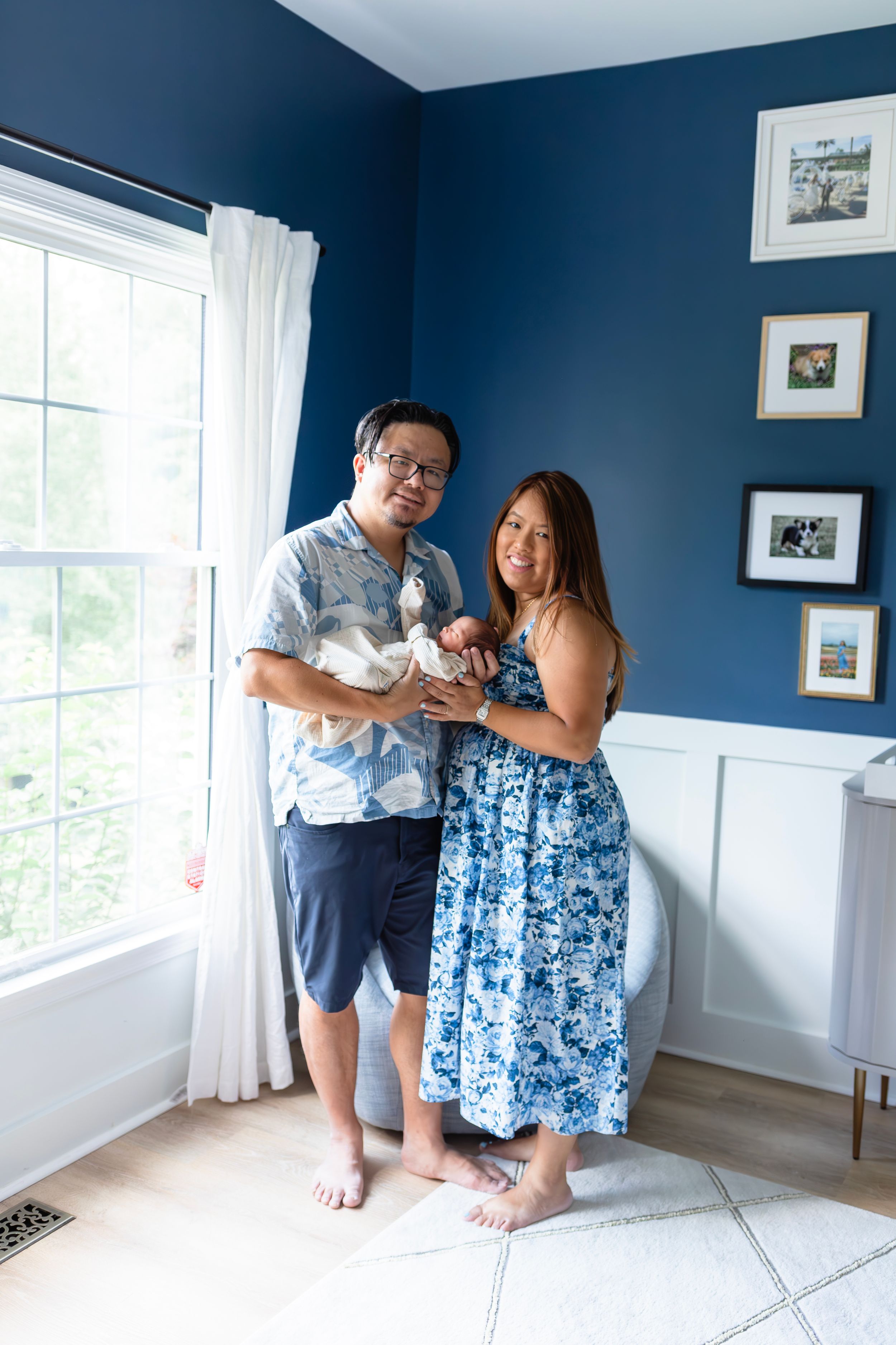 A couple stands in a blue nursery room holding their newborn baby.