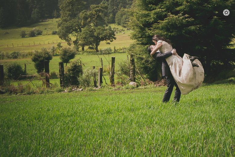 Groom lifts bride in flowing white dress on lush green meadow surrounded by trees and countryside.