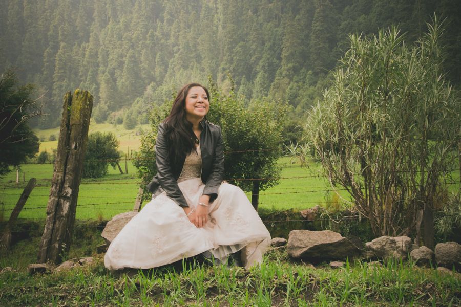 Smiling bride in white dress and leather jacket sits on rocks in scenic green forest setting.