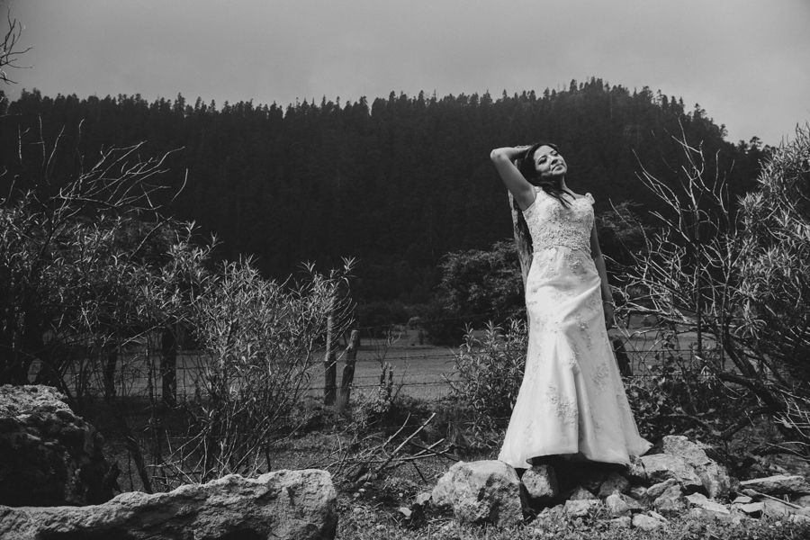 Elegant bride in fitted white wedding gown poses dramatically among trees in black and white portrait.