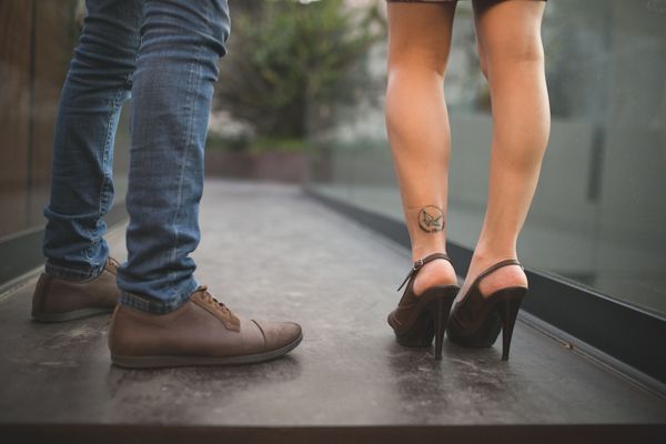 Close-up of couple standing close with woman in black heels showing rose ankle tattoo on pavement.