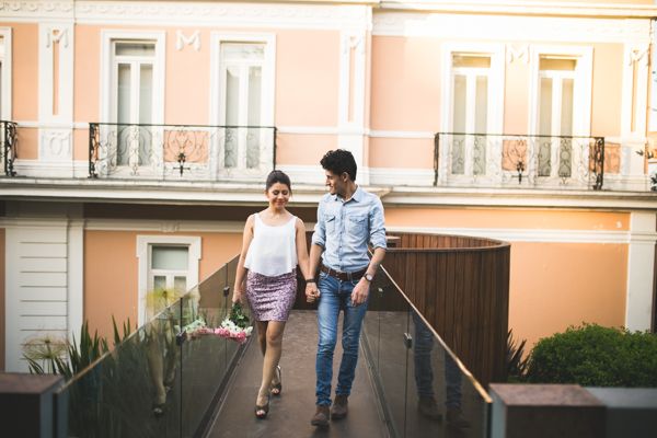 Happy couple walks hand in hand on terrace balcony in front of pink European-style building.