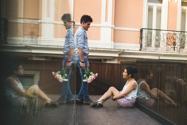 Couple in urban setting, man holding flowers approaching woman sitting against reflective glass wall.