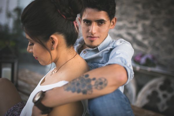 Young couple sitting back to back outdoors, man with tattoo on arm embracing woman in romantic pose.