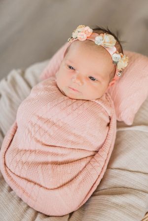 Newborn baby swaddled in soft pink knit blanket wearing delicate floral headband rests on cream colored fabric.