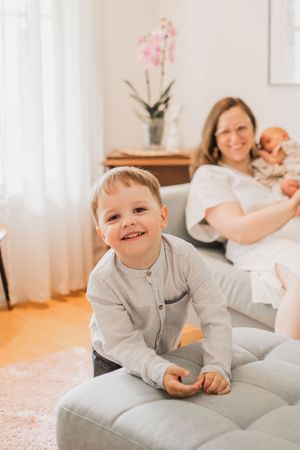 A joyful child in gray loungewear plays and smiles on a light-colored couch in a bright, airy room.