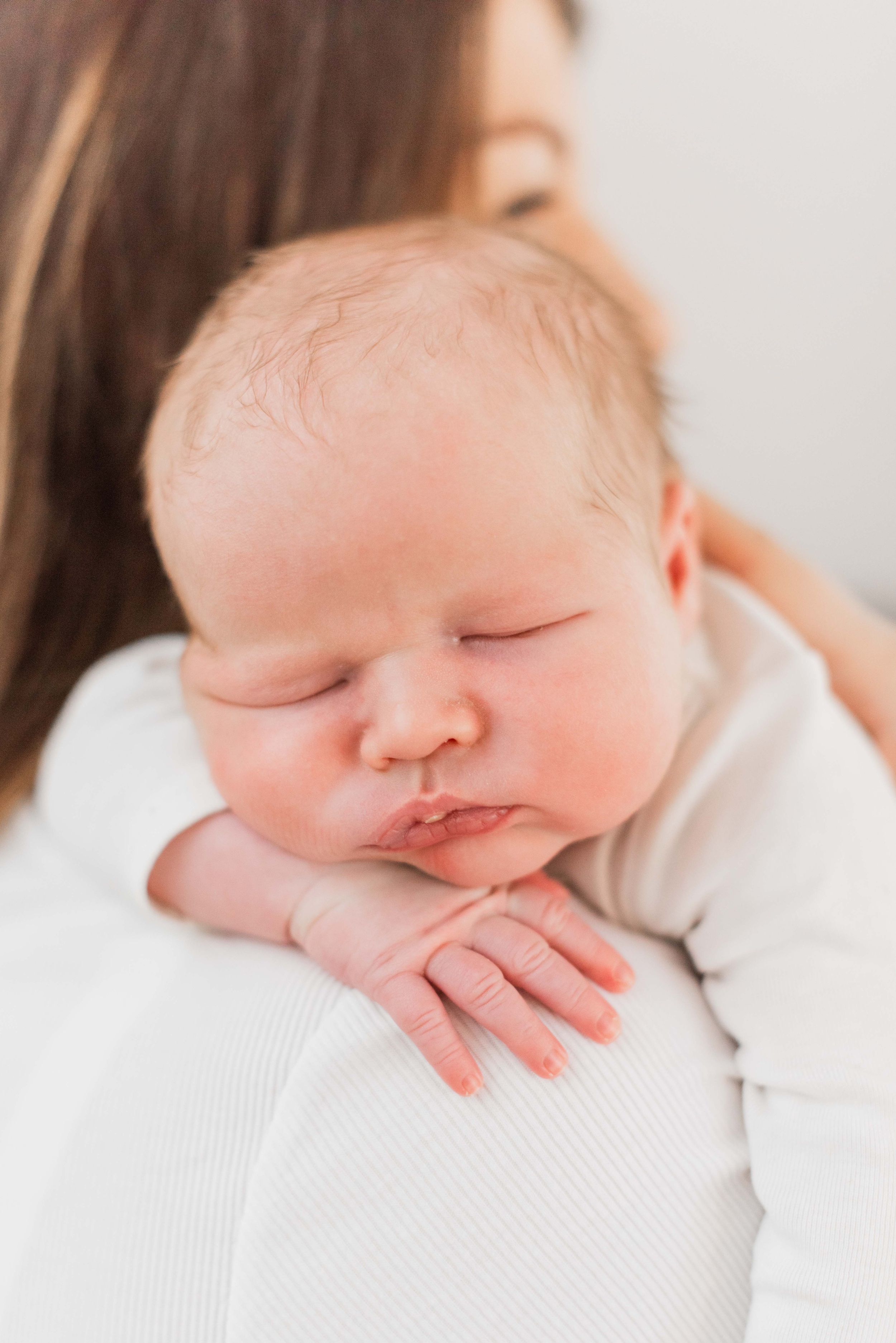 A sleeping newborn baby in white clothing rests peacefully on a white textured surface.