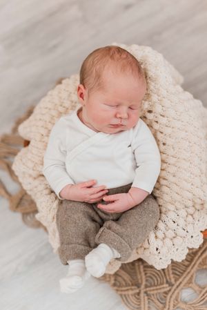 Sleeping newborn baby wearing white top and gray pants rests peacefully on cream knitted blanket.