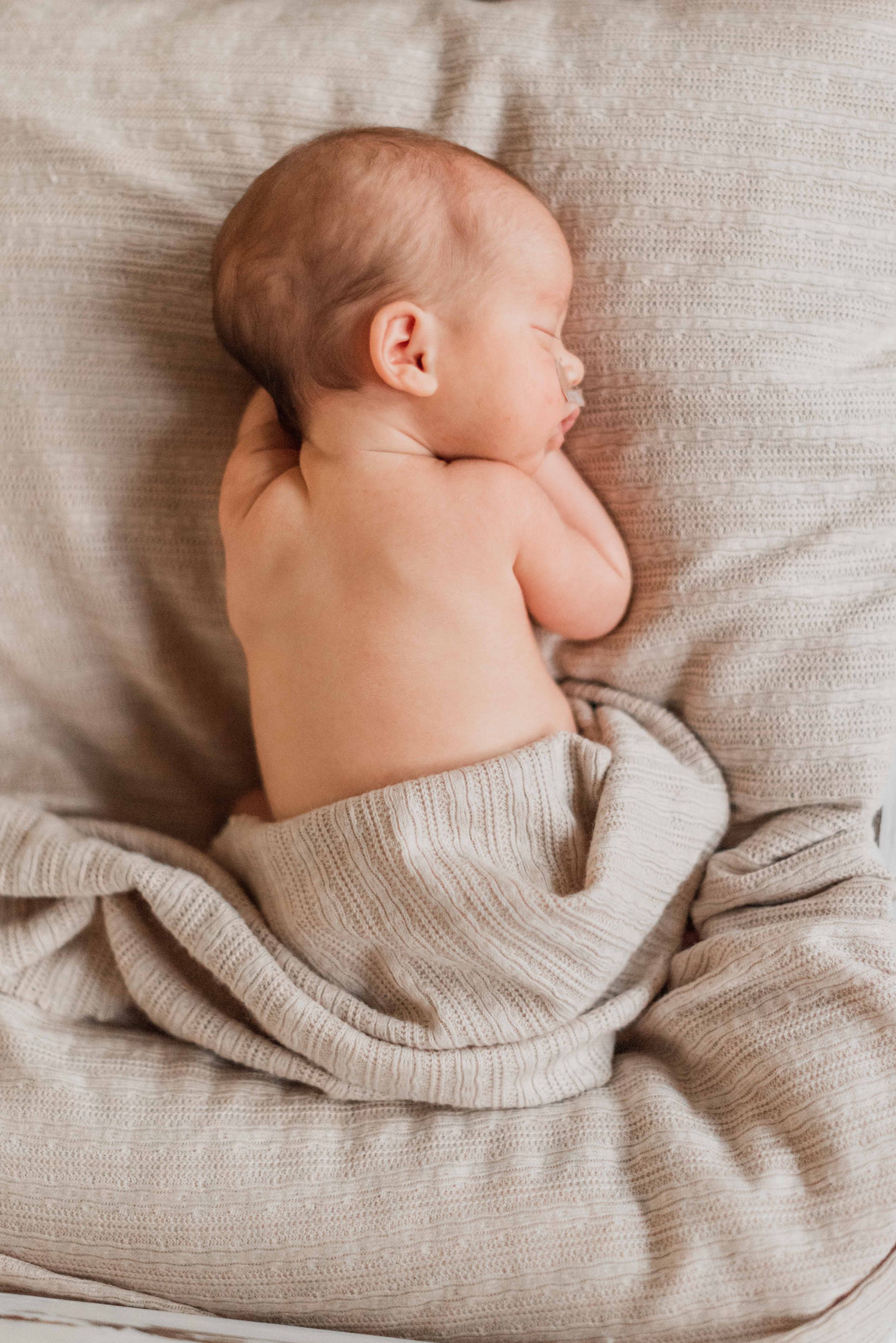 A sleeping newborn baby wrapped in a soft beige knit blanket lies on textured linen bedding.