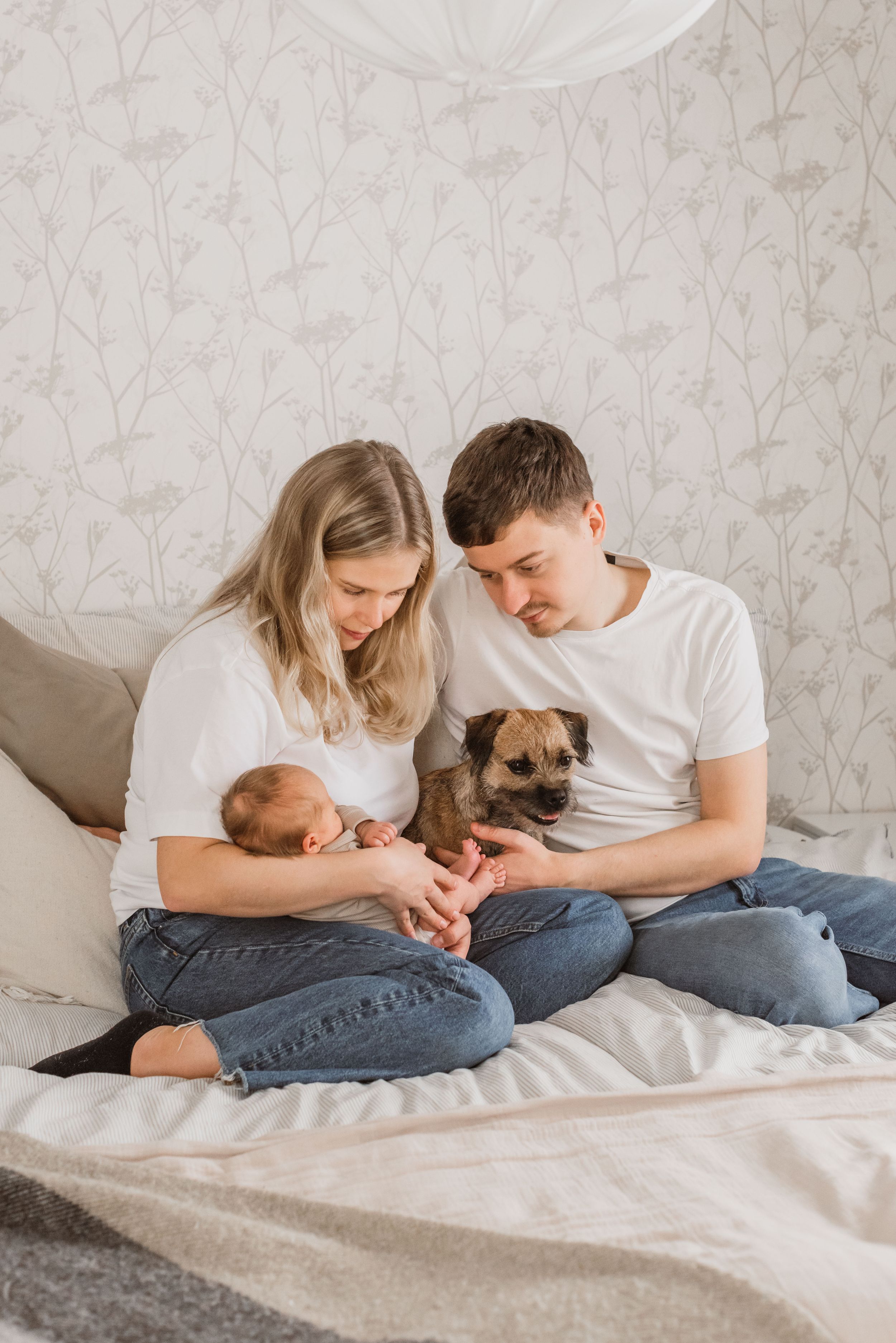 Young couple in white shirts and jeans sitting on bed with newborn baby and small brown dog in cozy bedroom setting.