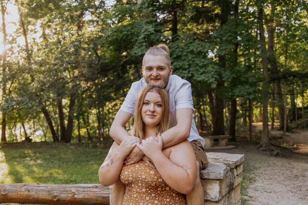 A loving couple poses together on a stone wall in a wooded setting.