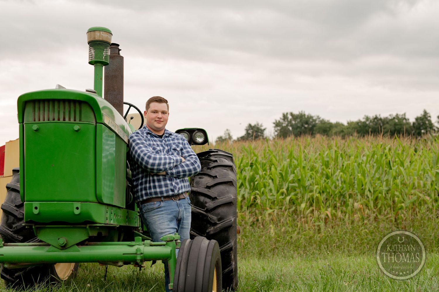 A person stands by the steering wheel of a classic John Deere tractor with cornfield in background.