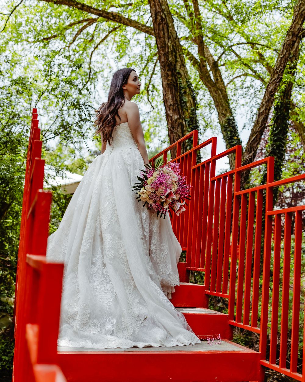 A person in wedding dress poses on vibrant red stairs surrounded by lush green trees.