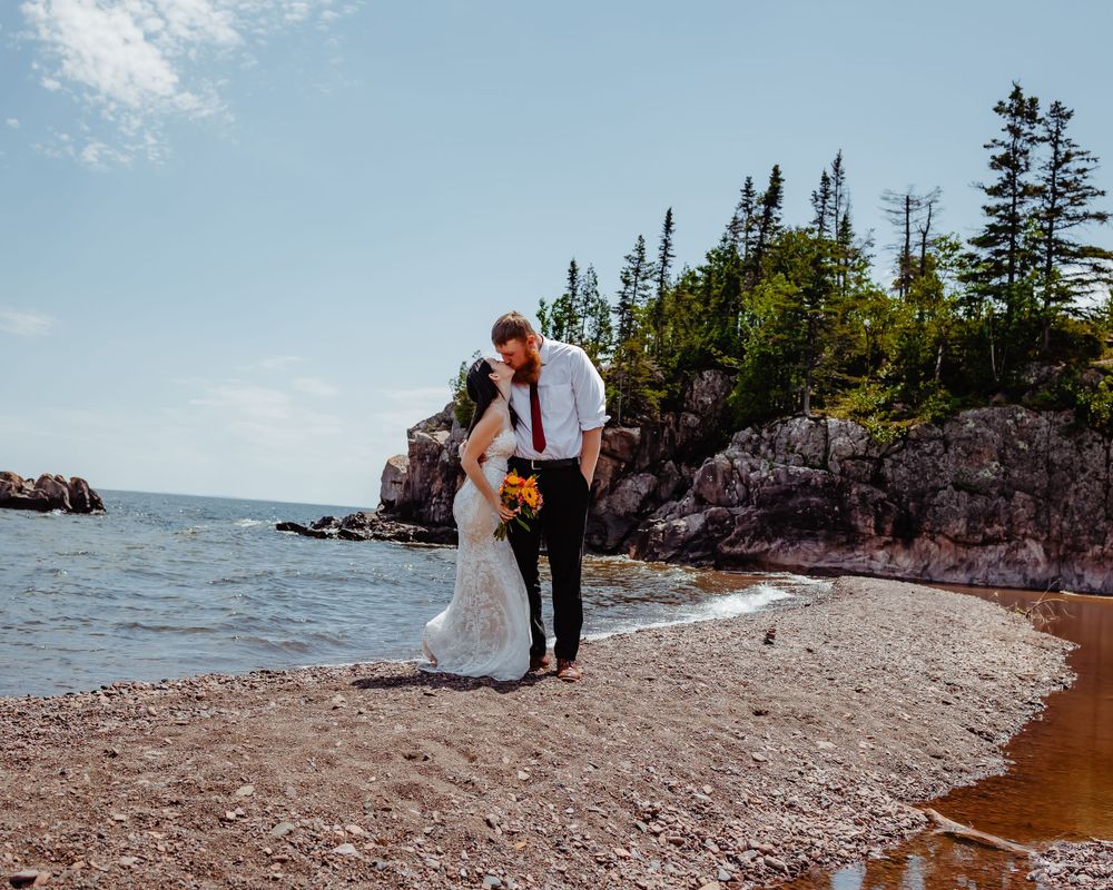 A couple shares a romantic moment on a rocky beach with evergreen trees and cliffs in the background.