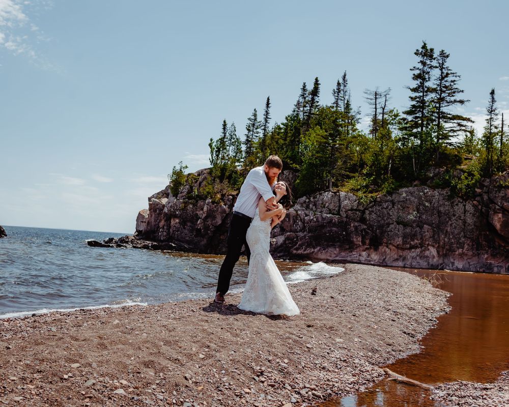 A couple embraces on a scenic Lake Superior shoreline with rocky cliffs and pine trees.