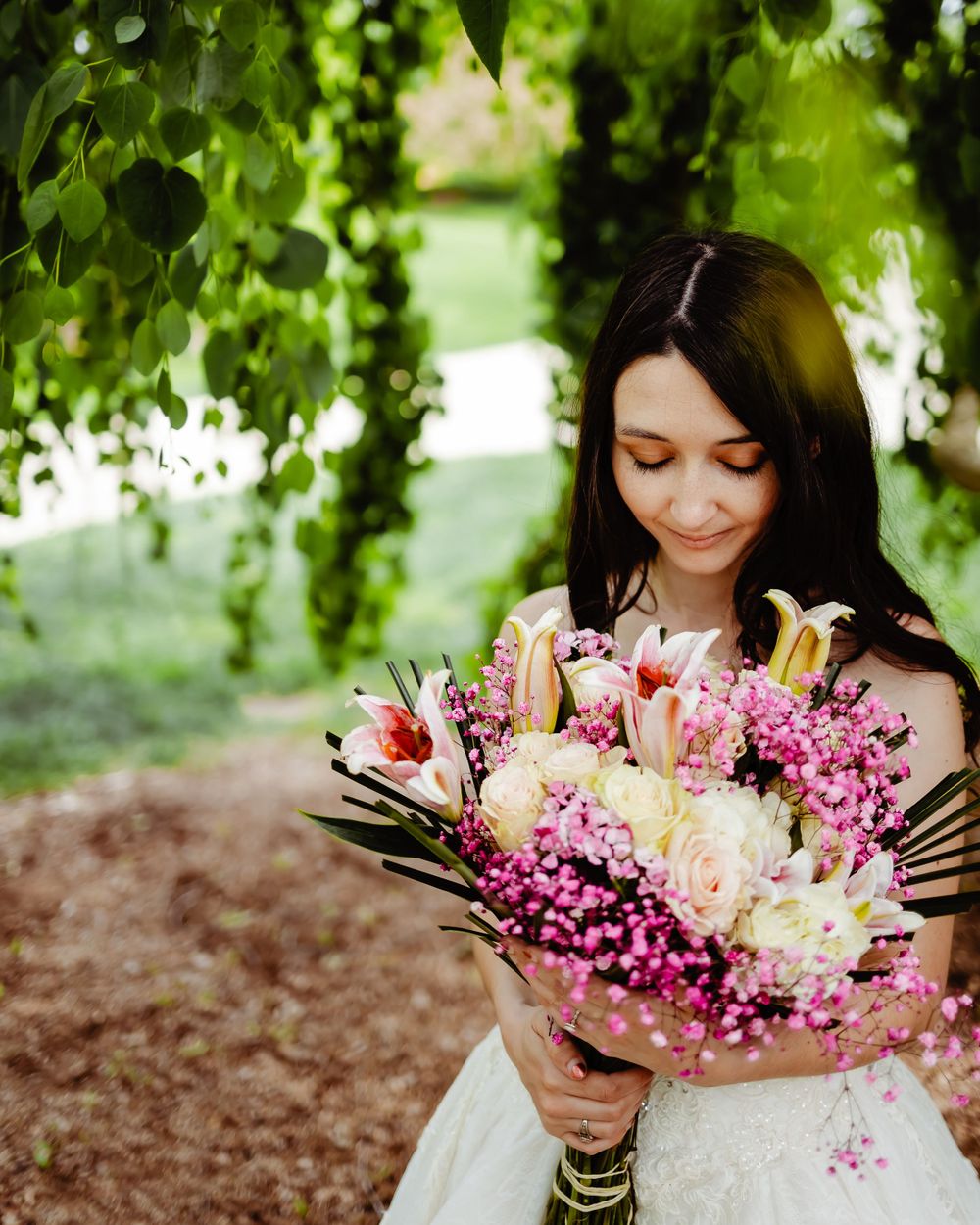 A person in white dress admires colorful bouquet of pink and white flowers under green foliage.