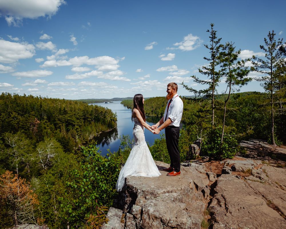 Couple in wedding attire poses on rocky overlook above winding river and forest.