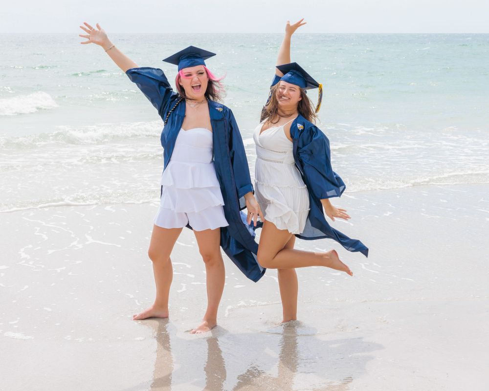 Two joyful graduates in blue caps and gowns dancing on a sunny white sand beach.