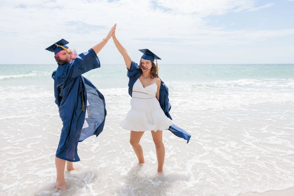Two graduates in blue caps and gowns giving high five while standing in ocean.