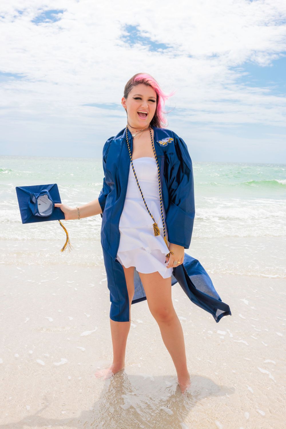 Excited graduate with pink hair in blue gown holding cap on a sunny beach.