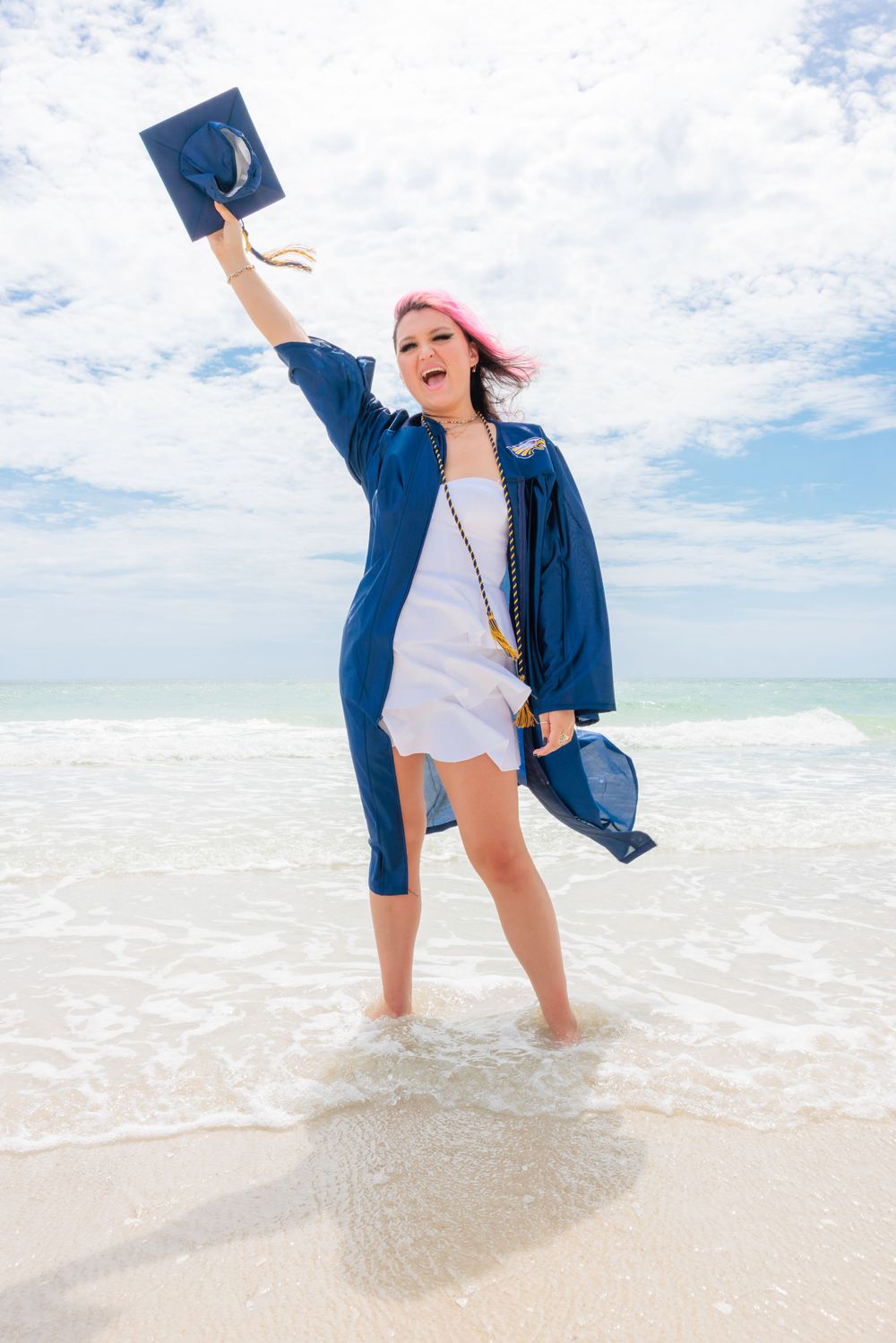 Excited graduate with pink hair celebrating in blue gown while standing in ocean waves.