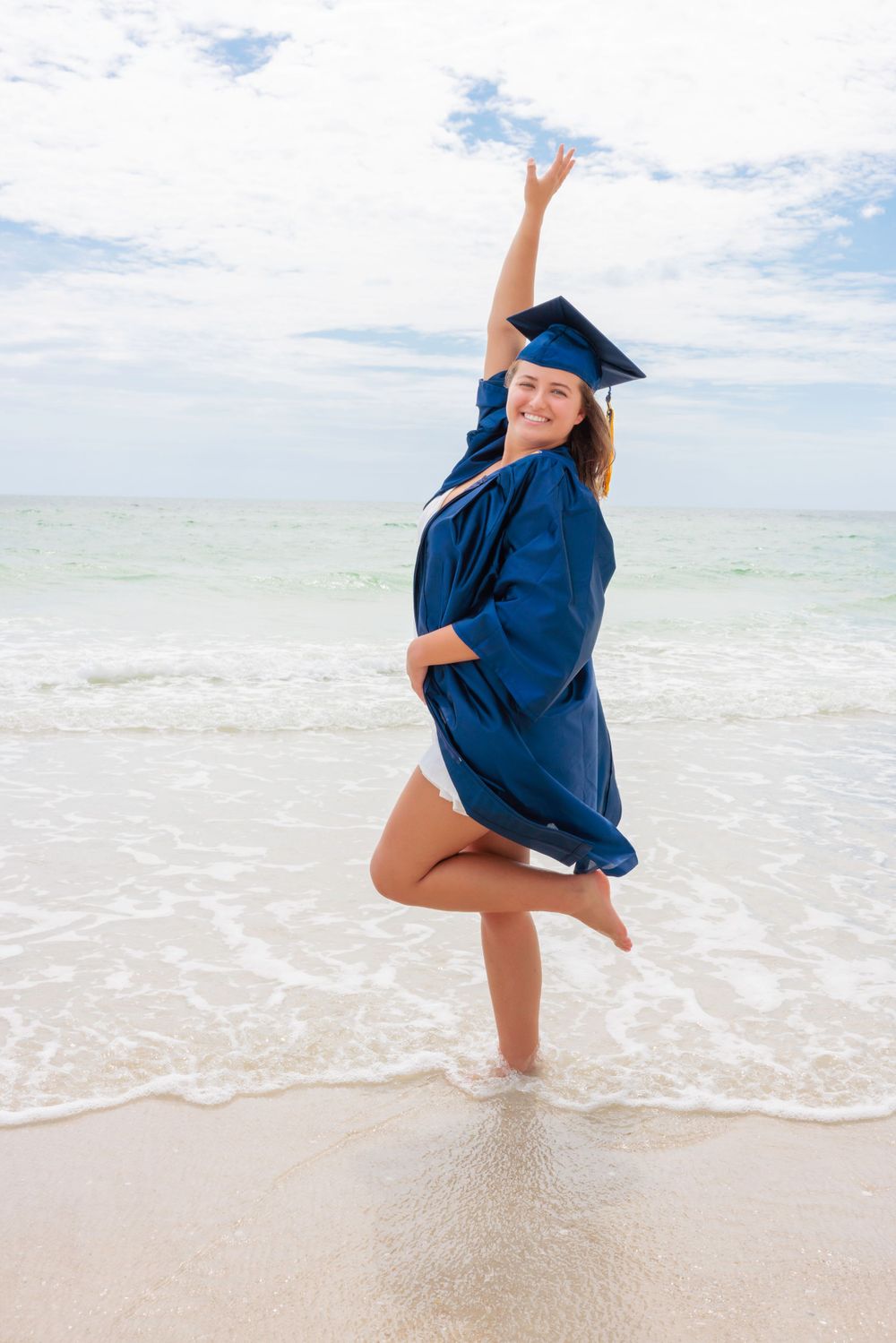 Celebrating graduate in blue cap and gown jumping joyfully on a sunny white sand beach.