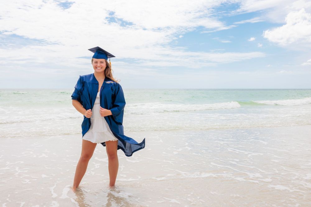 Smiling graduate in blue cap and gown walking through shallow ocean waves on a sunny beach.