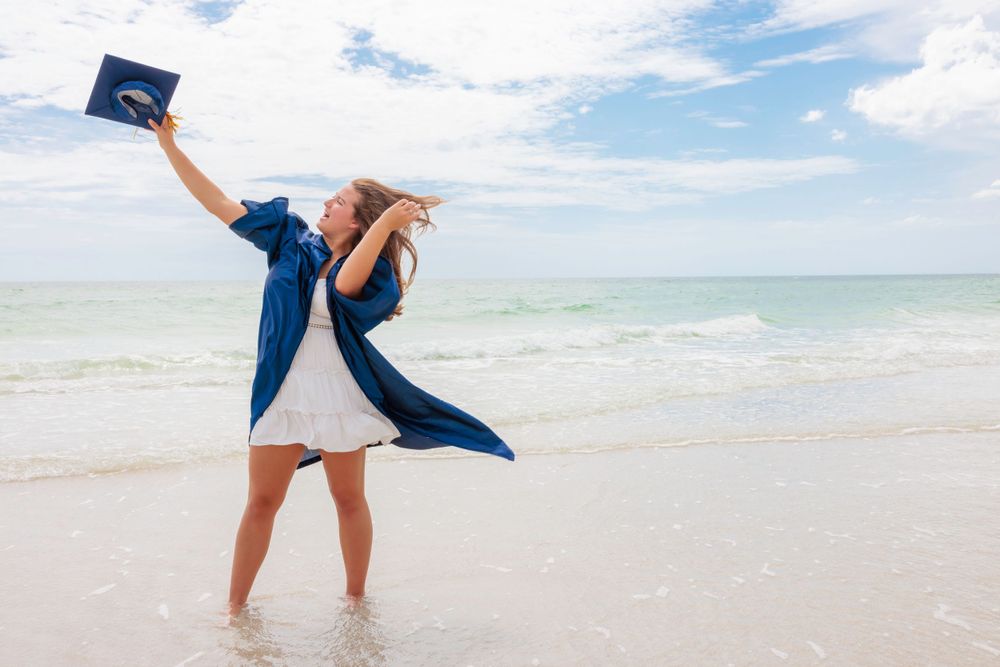 Excited graduate in blue gown raising cap triumphantly while standing in beach waves.