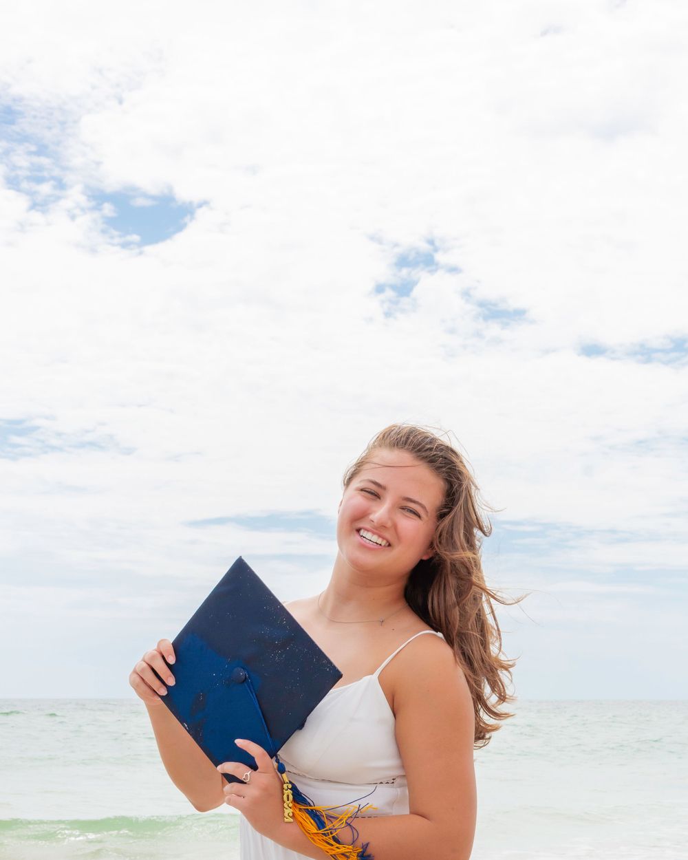 Smiling young woman holding blue graduation cap on a bright cloudy beach background.