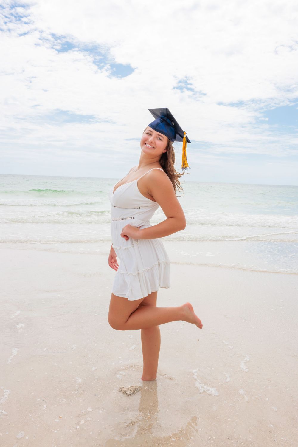 Smiling graduate in white dress and cap posing on a sunny beach with ocean waves.