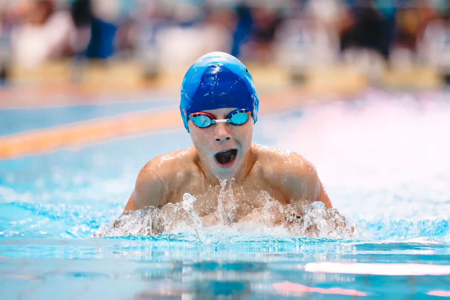 Swimmer emerges from water during breaststroke race wearing blue cap and goggles.