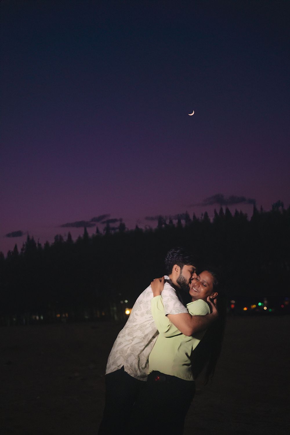 A couple embraces under a purple twilight sky with a crescent moon visible.