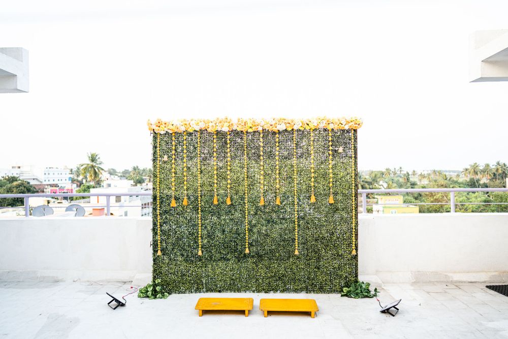 Green hedge wall backdrop decorated with hanging yellow tassels and marigold garlands for an Indian celebration.