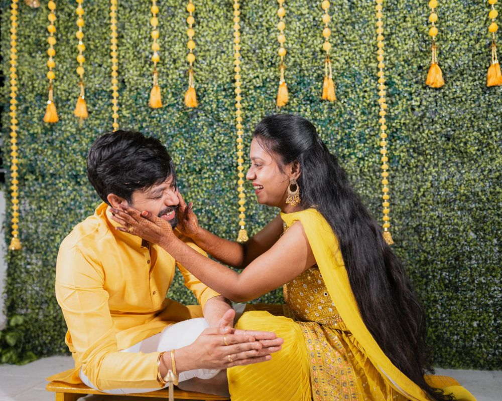 Couple in matching yellow outfits share playful moment against decorative green wall with hanging garlands.