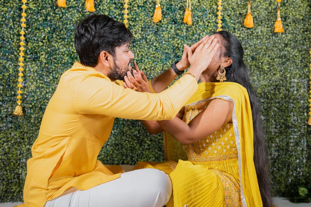 Joyful couple in yellow attire playing haldi ceremony games against floral backdrop.