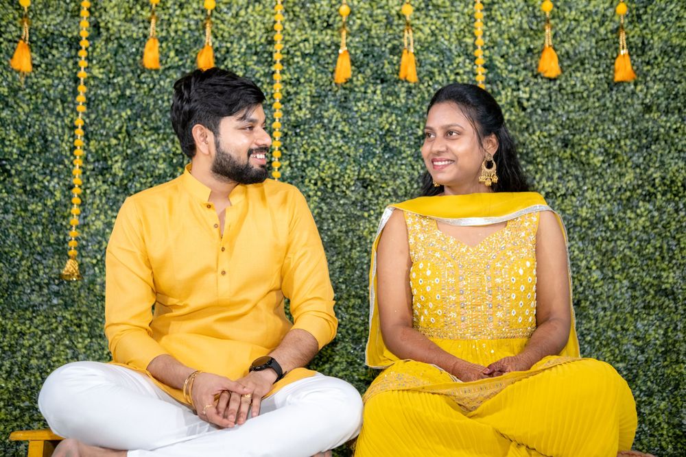 A couple in festive yellow Indian attire sits together against a green floral backdrop with marigold decorations.