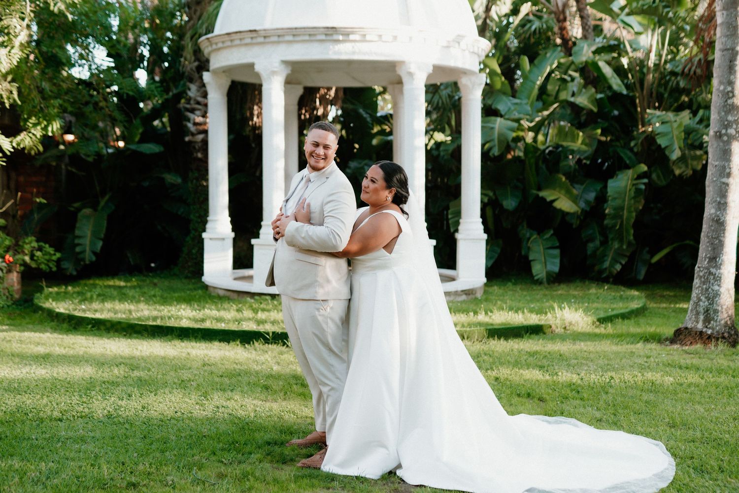 Newlyweds pose together under a white garden gazebo surrounded by lush tropical plants.