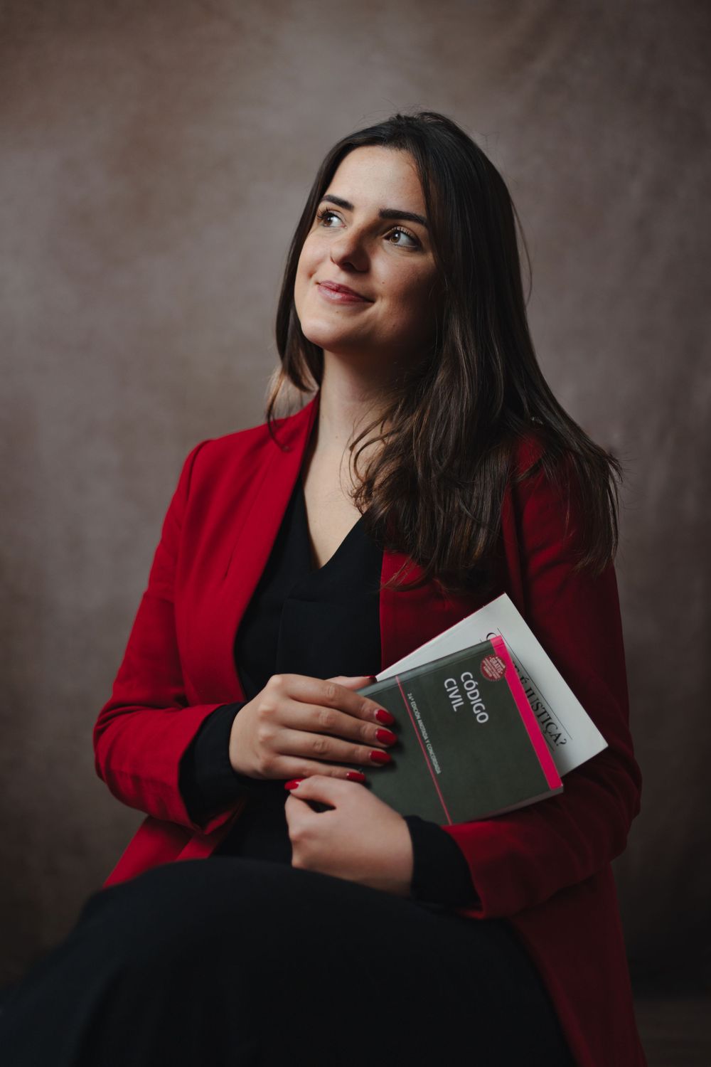 Professional portrait of person in red blazer and black top holding a book against dark background.