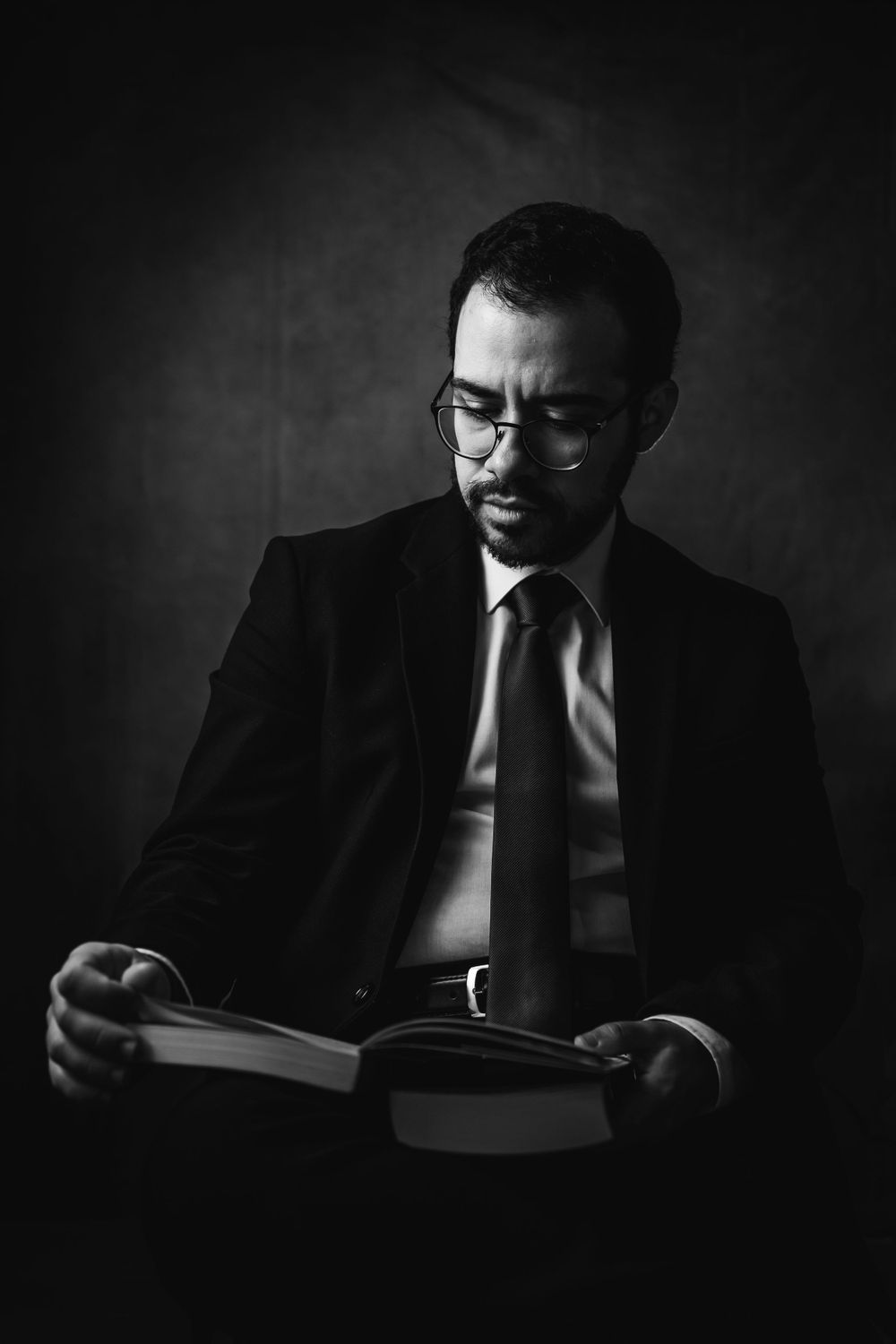 Professional black and white portrait of businessman in glasses reading book in dark setting.