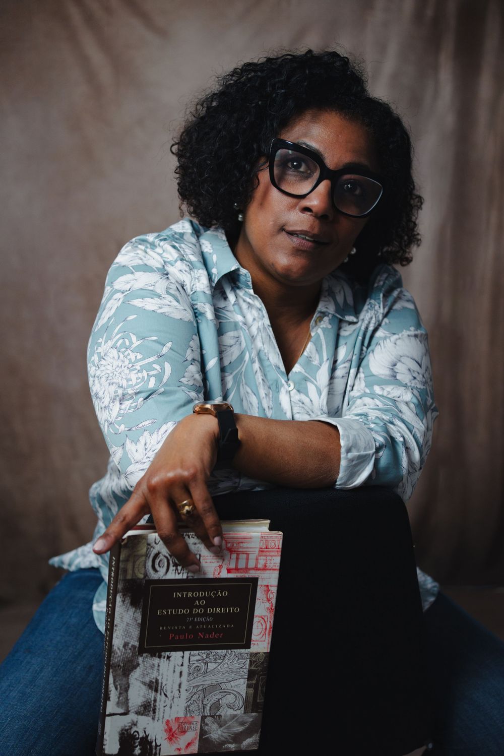 Portrait of person with curly hair and glasses holding book against neutral backdrop.