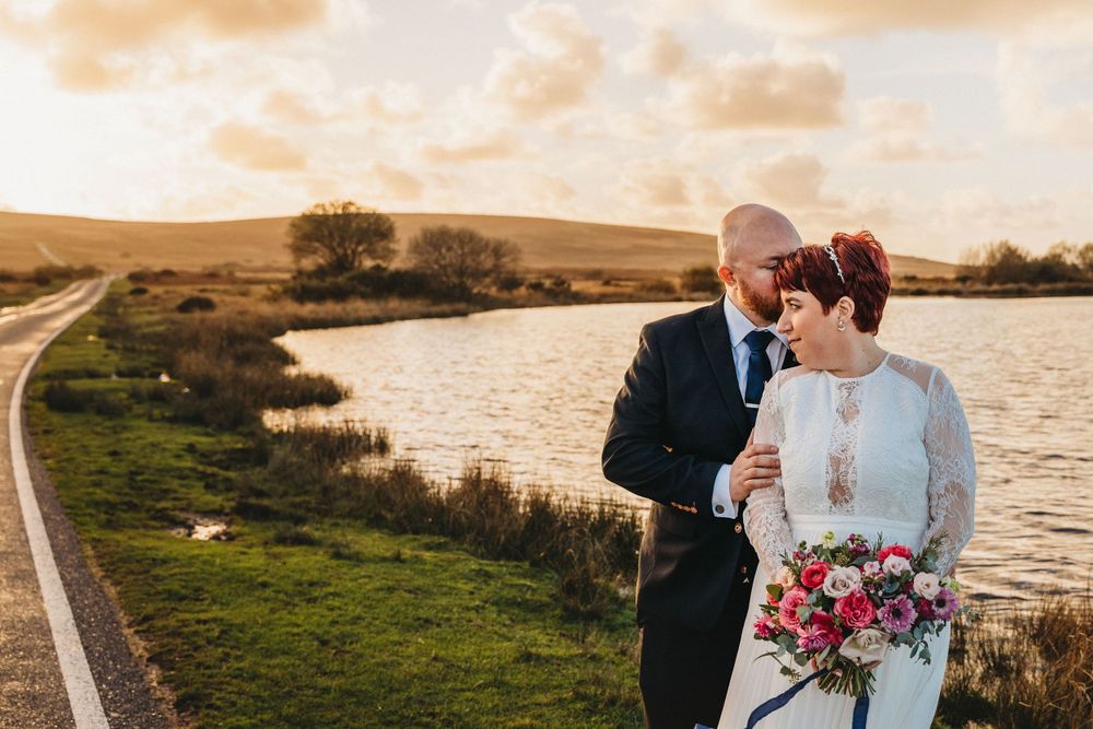 A couple shares an intimate moment by a lake during sunset while holding a colorful floral bouquet.