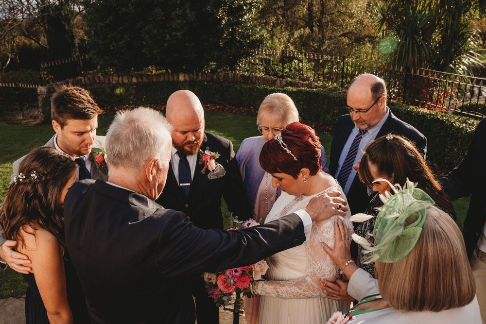Wedding guests gather in prayer circle during outdoor ceremony.