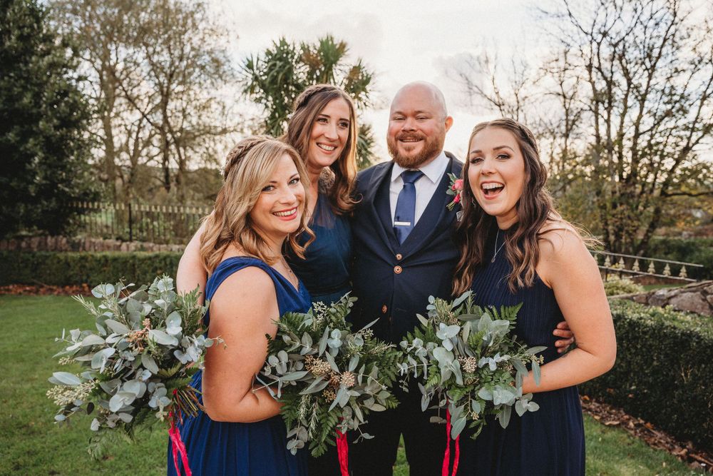 Wedding party in navy dresses and suits pose with greenery bouquets in an outdoor garden setting.