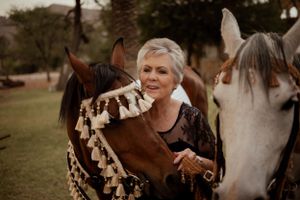 Woman with short gray hair affectionately pets two horses in an outdoor field setting.