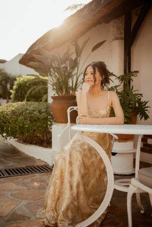 Woman in gold evening gown sits on white chair during golden hour.
