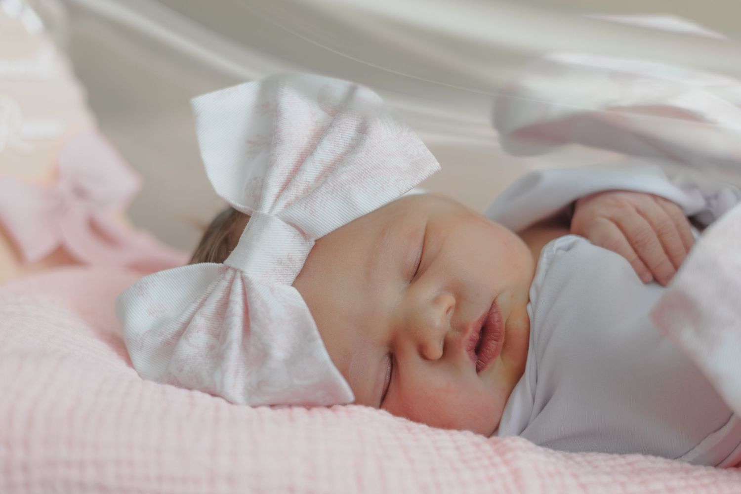 A newborn in pink and white striped clothing with a bow rests on white bedding.