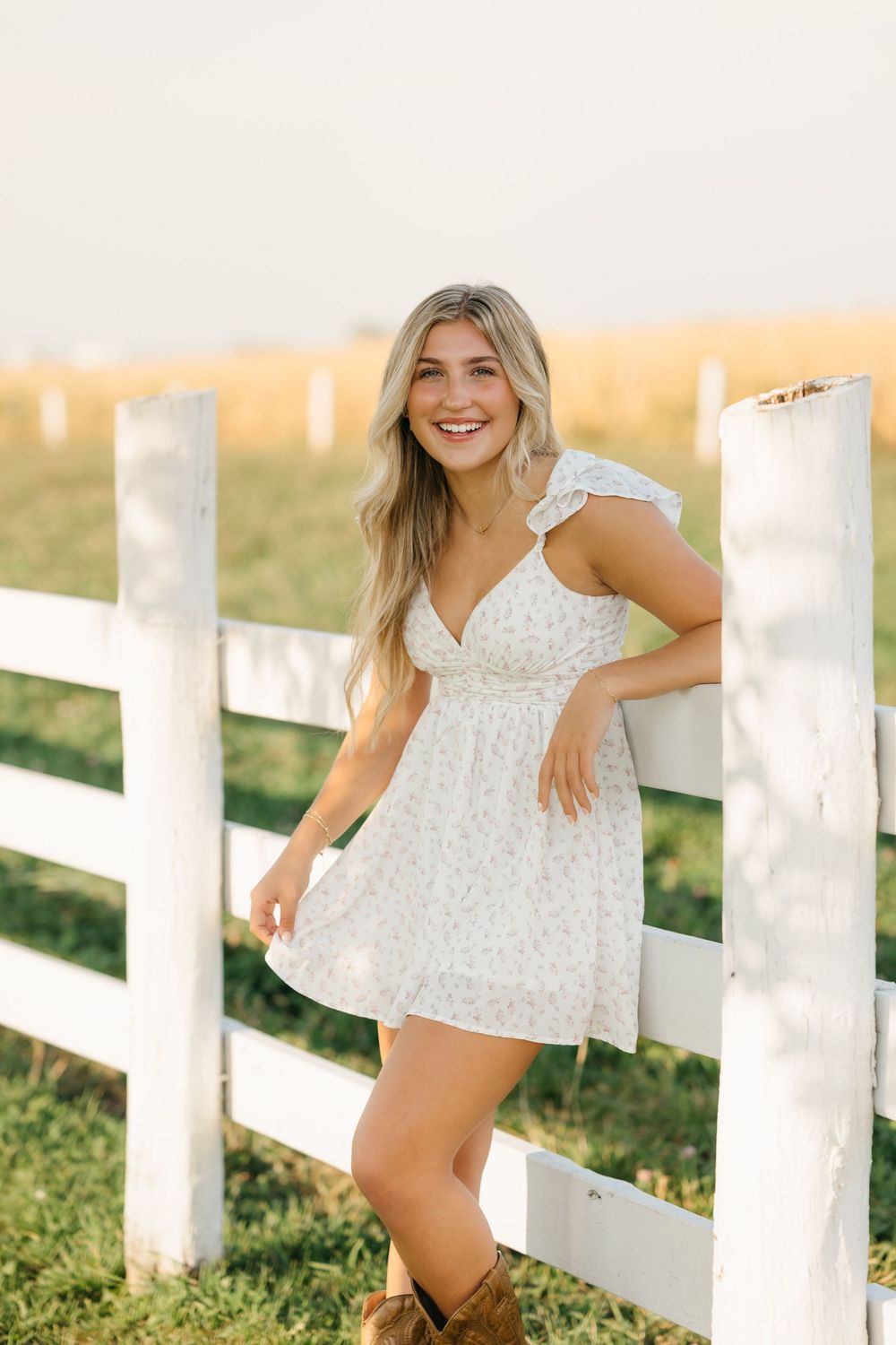 White floral sundress and brown boots against rustic white fence during golden hour sunset.