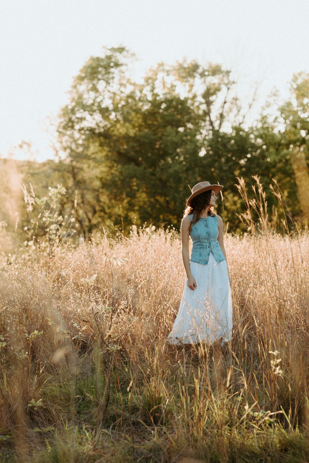 A person wearing a blue dress stands in a golden sunlit meadow of tall grass during sunset.