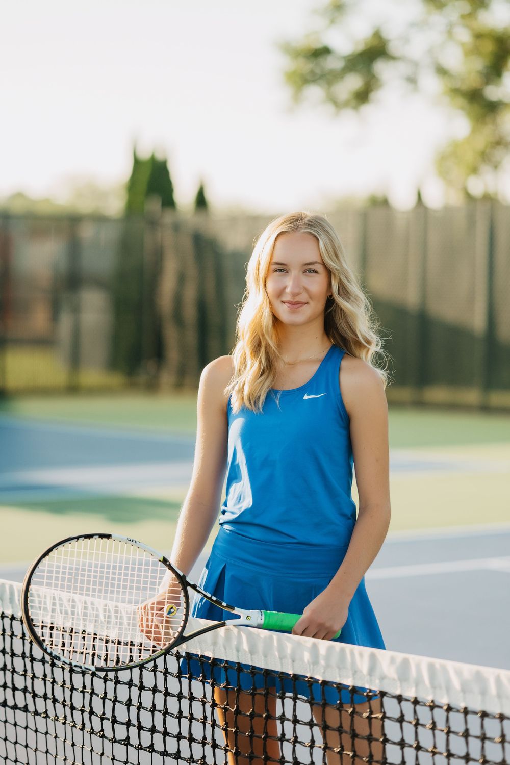 Tennis player wearing a blue Nike tank top and skirt stands smiling next to the court net with racquet in hand.